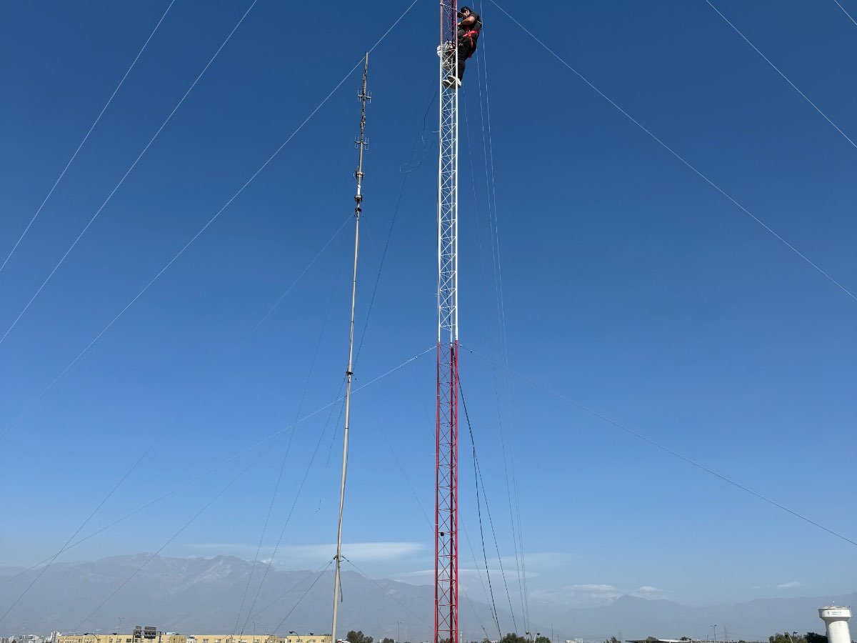Técnico trabajando en lo alto de una torre arriostrada roja y blanca, con un mástil de antenas al lado y la ciudad al fondo